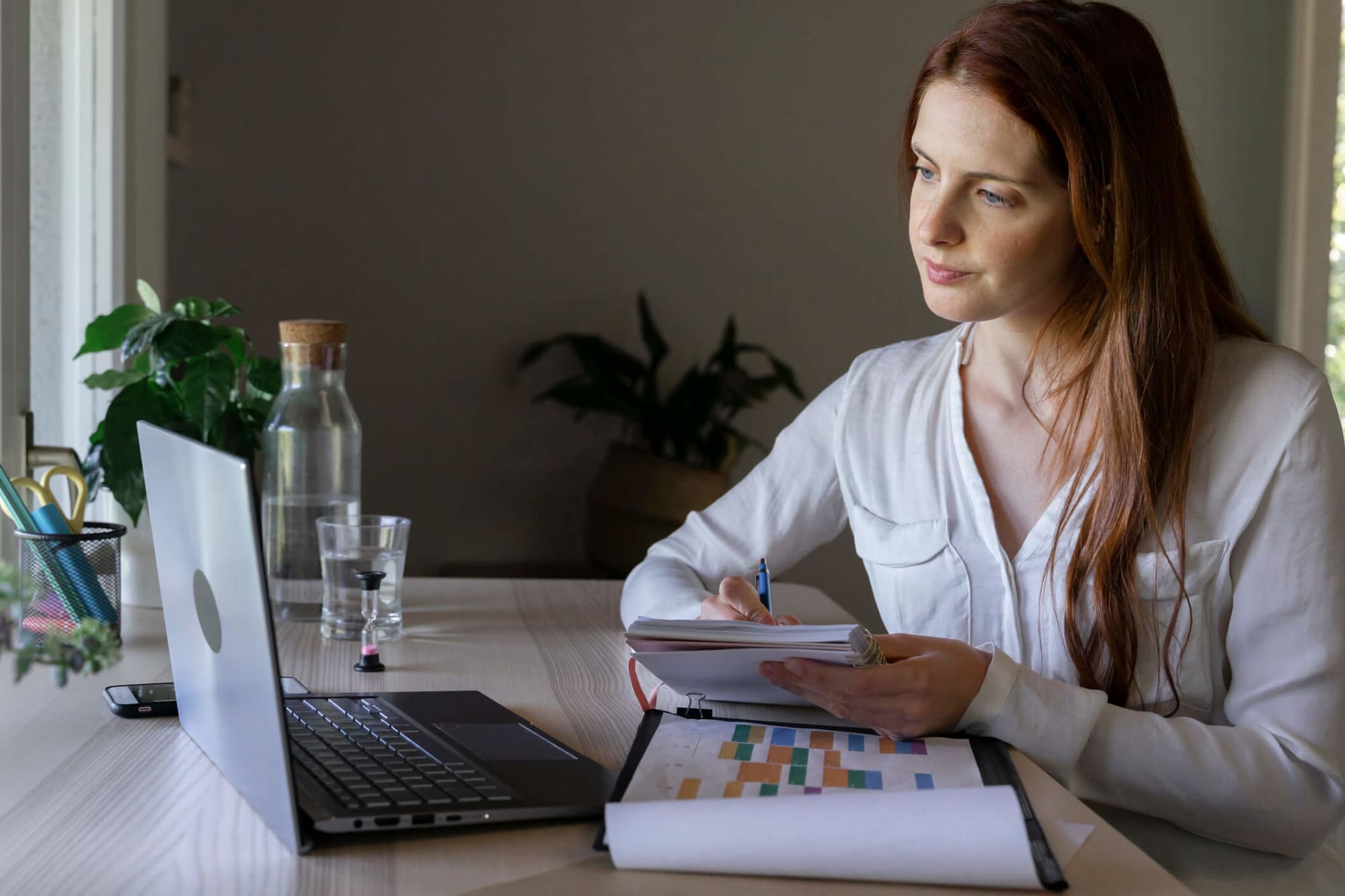 Psychologist writing notes in a book while working on her laptop at home.